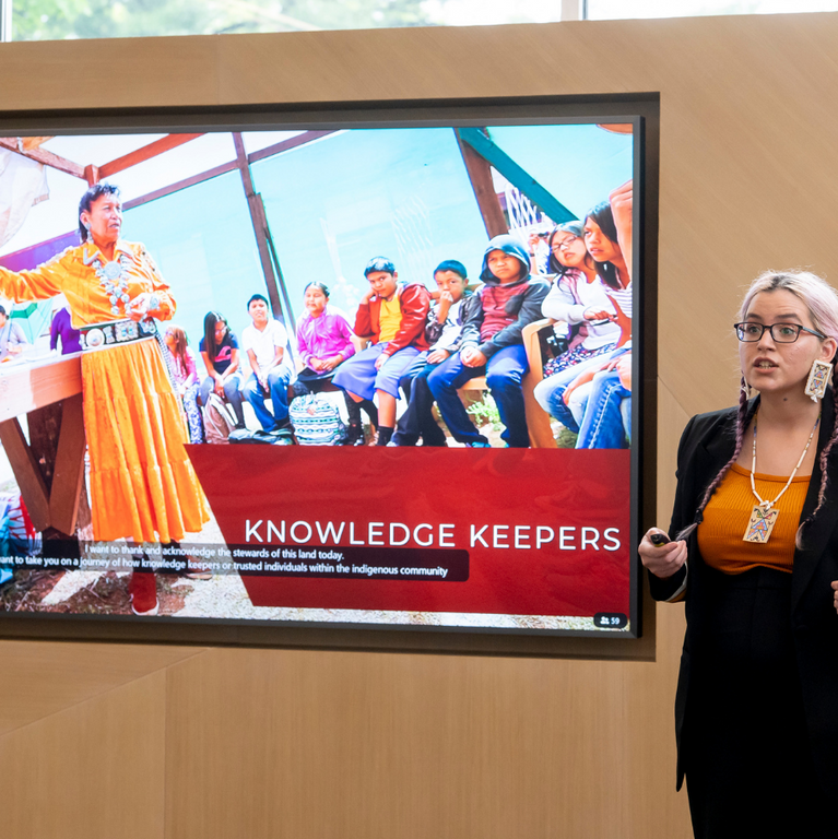 A woman delivers her pitch during the Empower Your Pitch 2023 contest standing next to her digital presentation which reads "Knowledge Keepers".