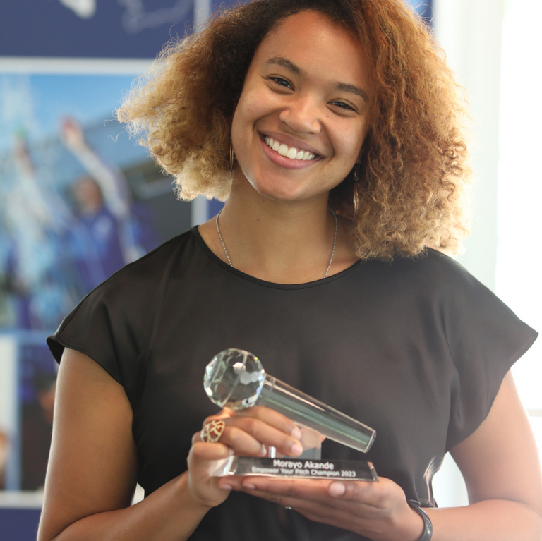 Empower Your Pitch 2023 winner Morayo Akande holds the Empower Your Pitch trophy, a glass microphone, and smiles.